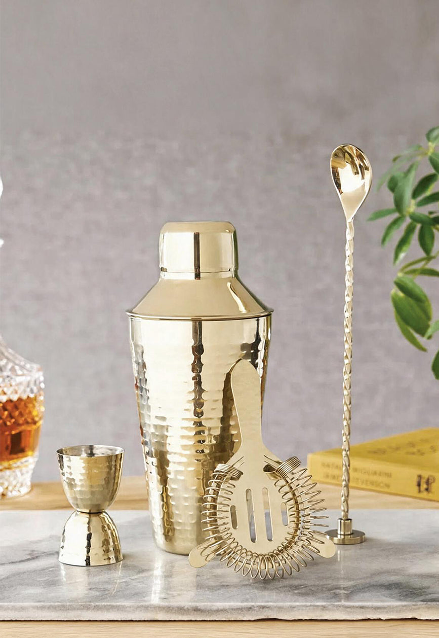 Hammered gold cocktail shaker with jigger, Hawthorne strainer and twisted bar spoon on marble