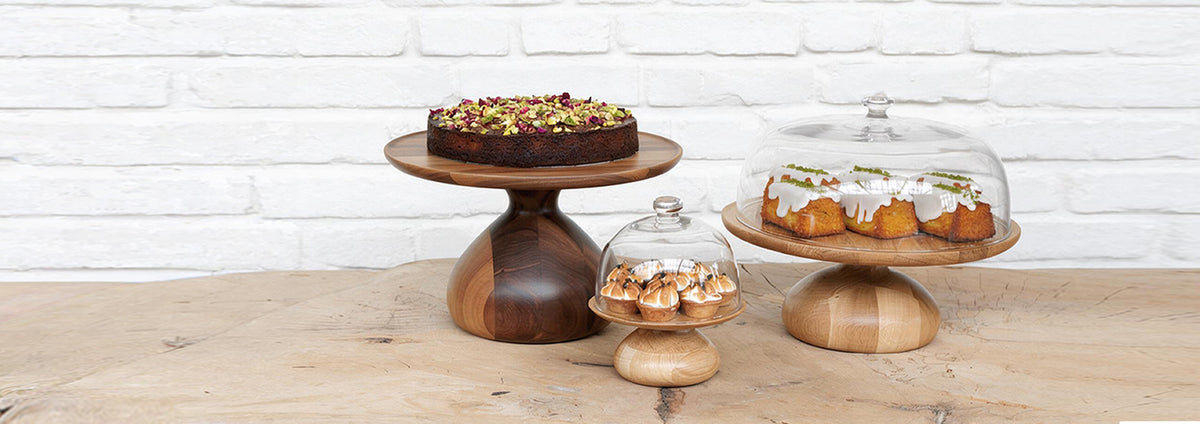 Walnut and oak cake stands with cake, pastries and glass domes on a rustic wooden table against a white brick wall