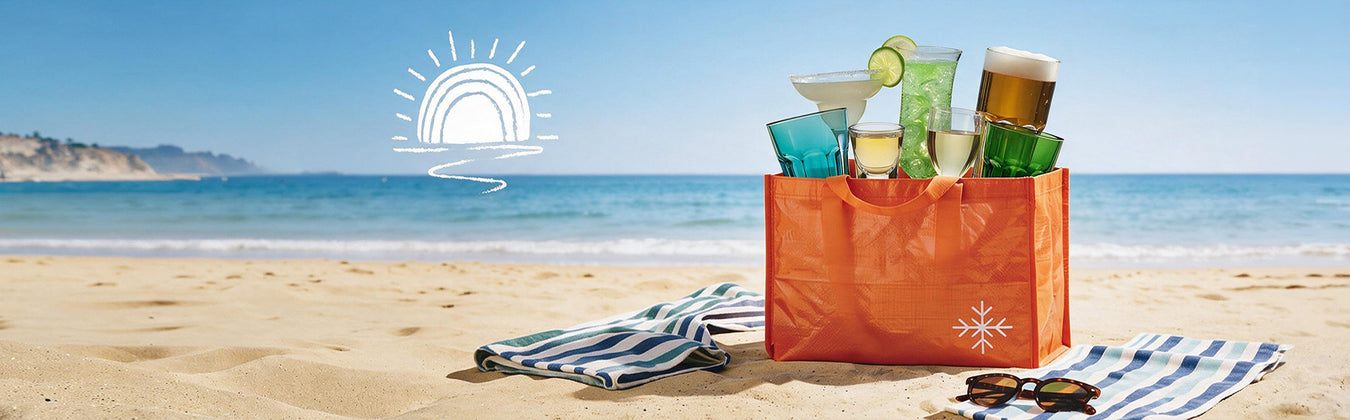 An orange Restofair beach tote on the sand, filled with summer drinkware — a margarita coupe, shot glass, frozen cocktail, champagne flute, beer glass and coloured tumblers — beside striped towels and sunglasses, with the sea behind